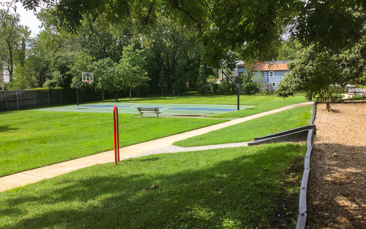 Outdoor park with two basketball courts, a bench, walking paths, grassy area, trees, and a playground area with wood chips in the foreground.