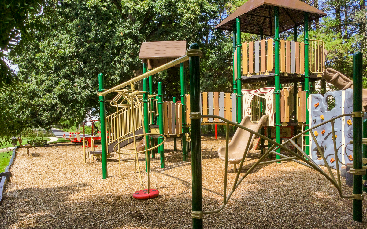 Outdoor playground with slides, climbing structures, and a swing set on a wood chip surface, surrounded by trees and greenery.