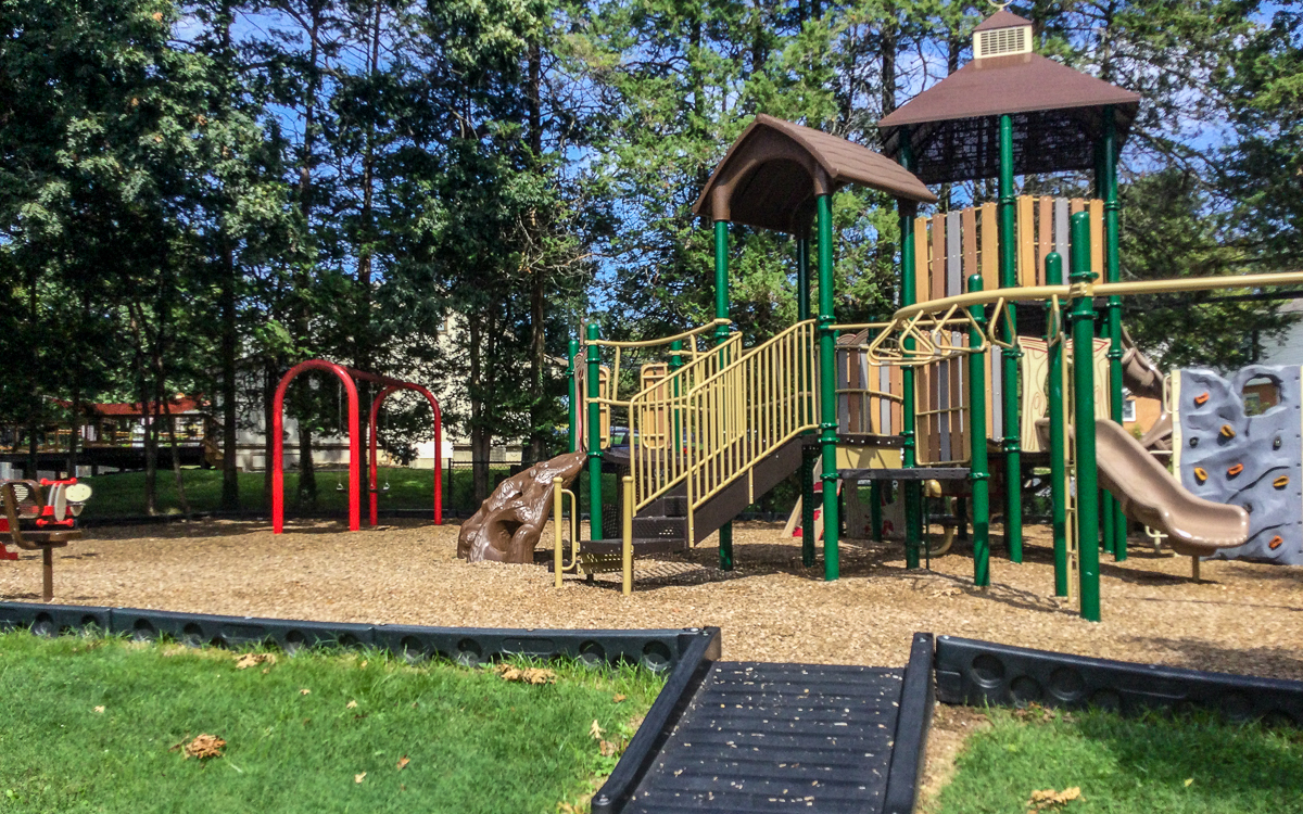 Outdoor playground with climbing structures, slides, a bridge, and swings, surrounded by trees and wood chip ground cover.
