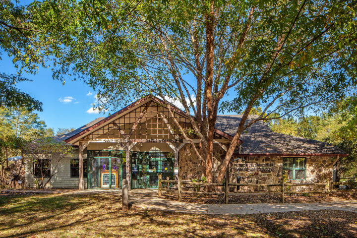 Single-story stone building with large windows and a peaked roof, surrounded by trees and a wooden fence, under a clear blue sky.