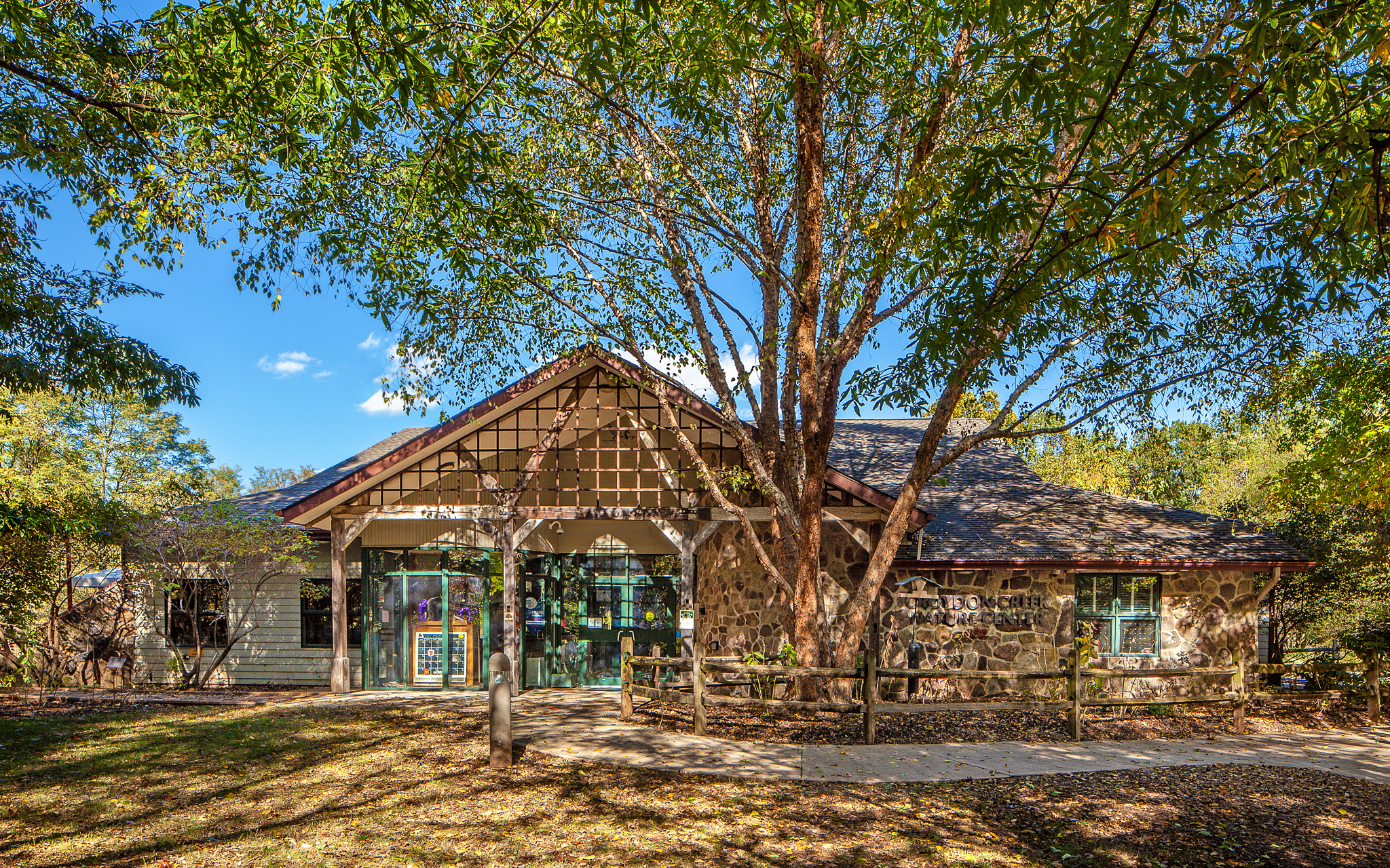 Single-story stone building with large windows and a peaked roof, surrounded by trees and a wooden fence, under a clear blue sky.