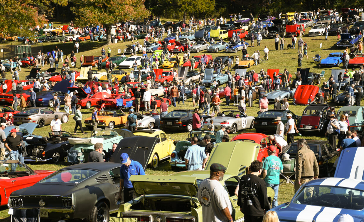 A large outdoor car show with many classic cars on display, open hoods, and crowds of people walking among the vehicles on a sunny day.