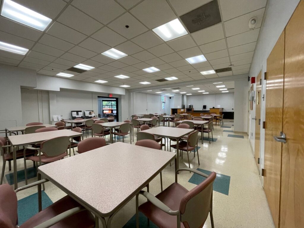 A well-lit cafeteria with empty tables and chairs, vending machines in the back, and a counter area along the far wall.