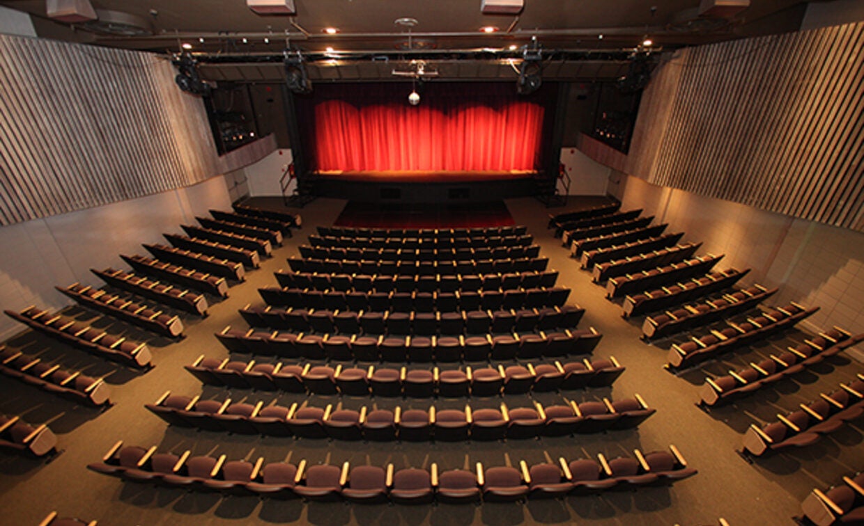 An empty auditorium with rows of seats facing a stage with a closed red curtain, viewed from the back of the room.