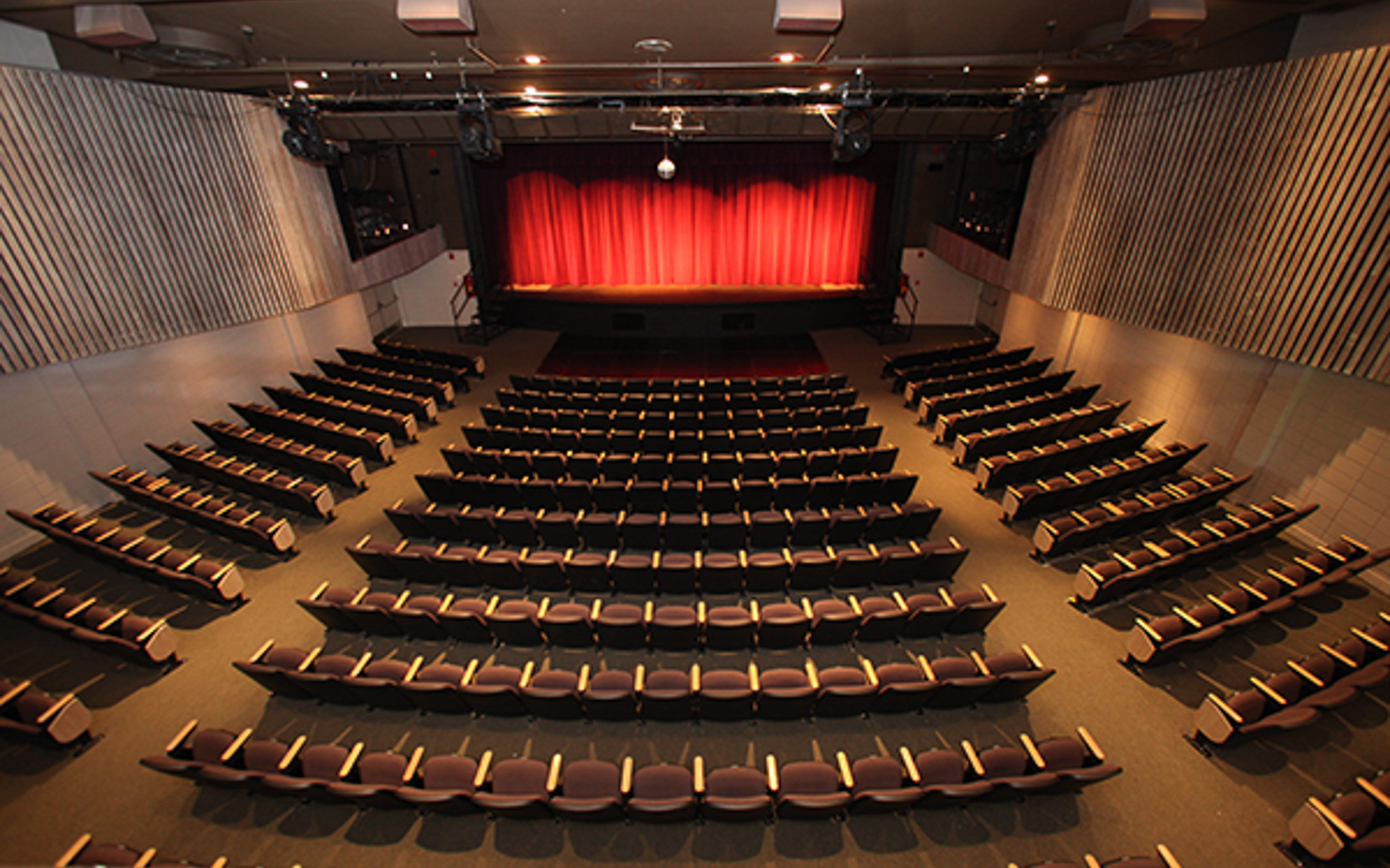 An empty auditorium with rows of seats facing a stage with a closed red curtain, viewed from the back of the room.