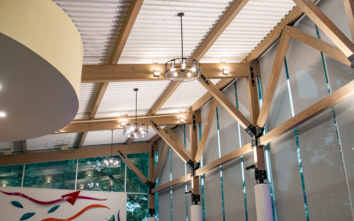 Interior view of a modern building with exposed wooden beams, large windows with blinds, circular light fixtures, and abstract wall art.