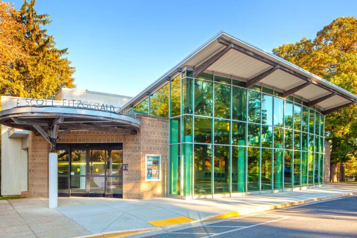 Modern building with large glass windows and an overhang at the entrance, surrounded by trees and bathed in sunlight. The sign reads 
