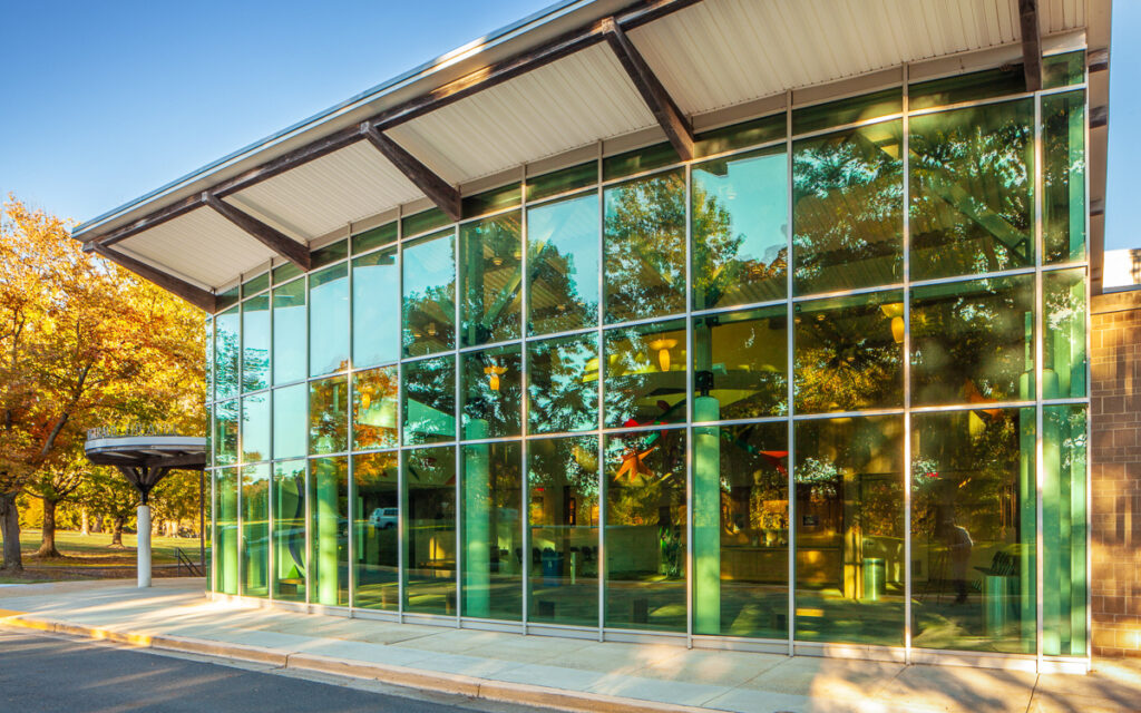 A modern building with large glass windows reflecting trees and a clear sky, with a sidewalk and paved area in front.
