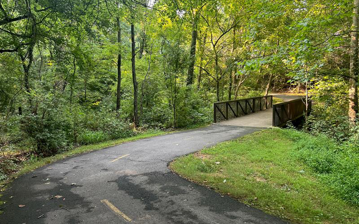 A paved walking trail curves through a forested area and crosses a small wooden bridge surrounded by green trees and grass.