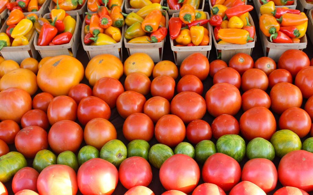 Rows of orange, red, and green heirloom tomatoes are arranged in front of baskets filled with yellow and red mini bell peppers.