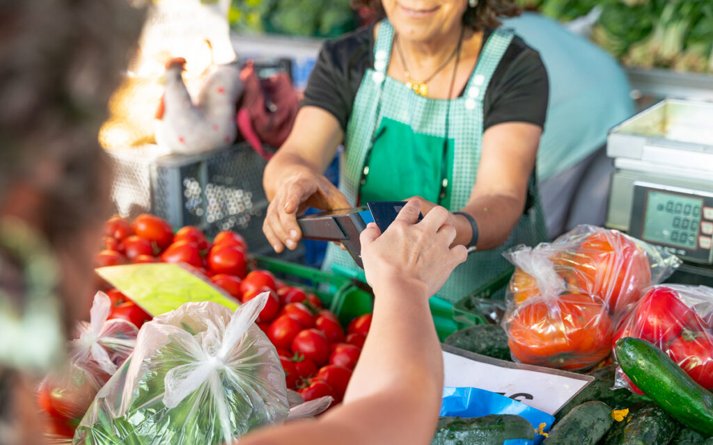 A vendor in a green apron accepts a card payment from a customer at a market stall with tomatoes, peppers, and other vegetables.