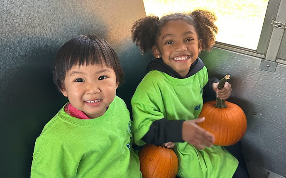 Two young children in green shirts sit on a bench holding small pumpkins and smiling at the camera.