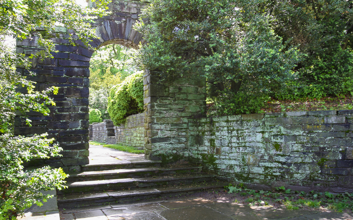 Stone archway and steps surrounded by lush greenery and moss-covered stone walls in an outdoor garden setting.