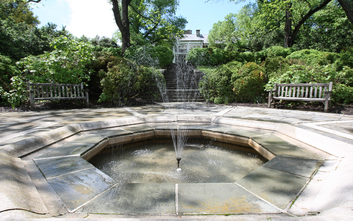 A small circular fountain with water jets in a stone-paved area, flanked by two benches, surrounded by green bushes and trees, with a building visible in the background.