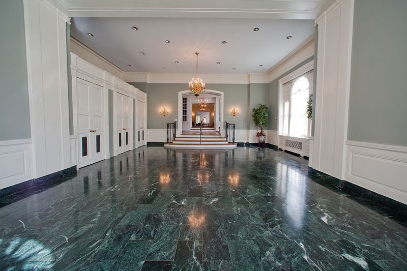 Spacious hallway with green marble floor, white paneled walls, large windows, and a chandelier above a small staircase leading to double doors.