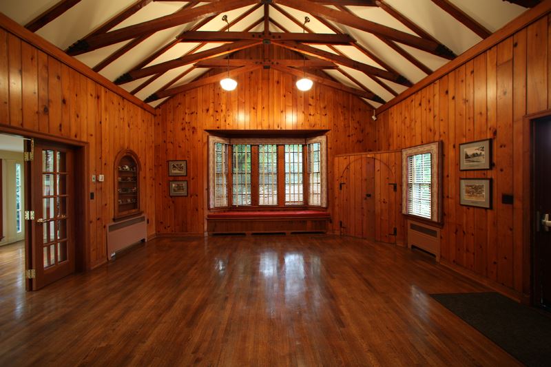 A spacious, empty room with wood-paneled walls, hardwood floors, exposed ceiling beams, and large windows letting in natural light.
