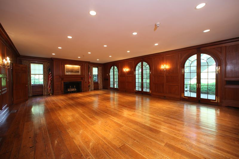 Spacious, empty room with wooden floors, wood-paneled walls, arched windows, recessed ceiling lights, fireplace, and American flags by the windows.