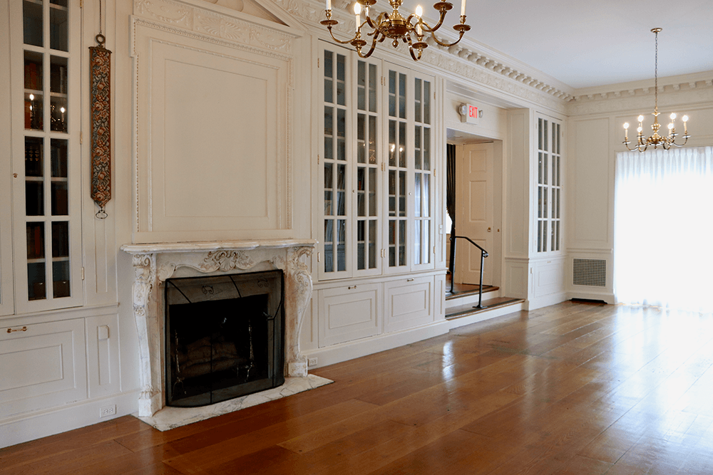 A bright, empty room with wooden floors, a white marble fireplace, built-in glass cabinets, chandeliers, and large windows with sheer curtains.
