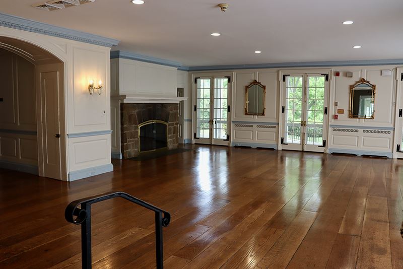 A spacious, empty room with hardwood floors, a stone fireplace, wall mirrors, and large windows letting in natural light.