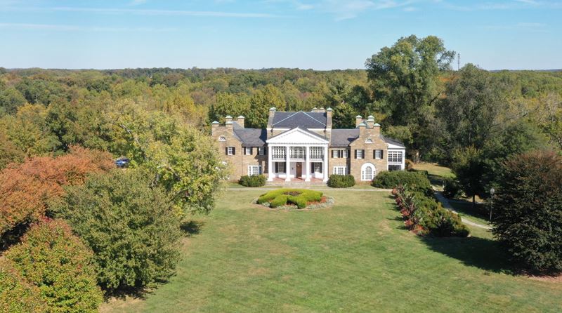 A large two-story stone mansion with white columns stands surrounded by trees and landscaped lawns under a clear blue sky.