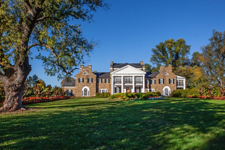 Large two-story mansion with brick exterior, white columns, and multiple chimneys, set on a landscaped lawn with trees and colorful flowerbeds under a clear blue sky.