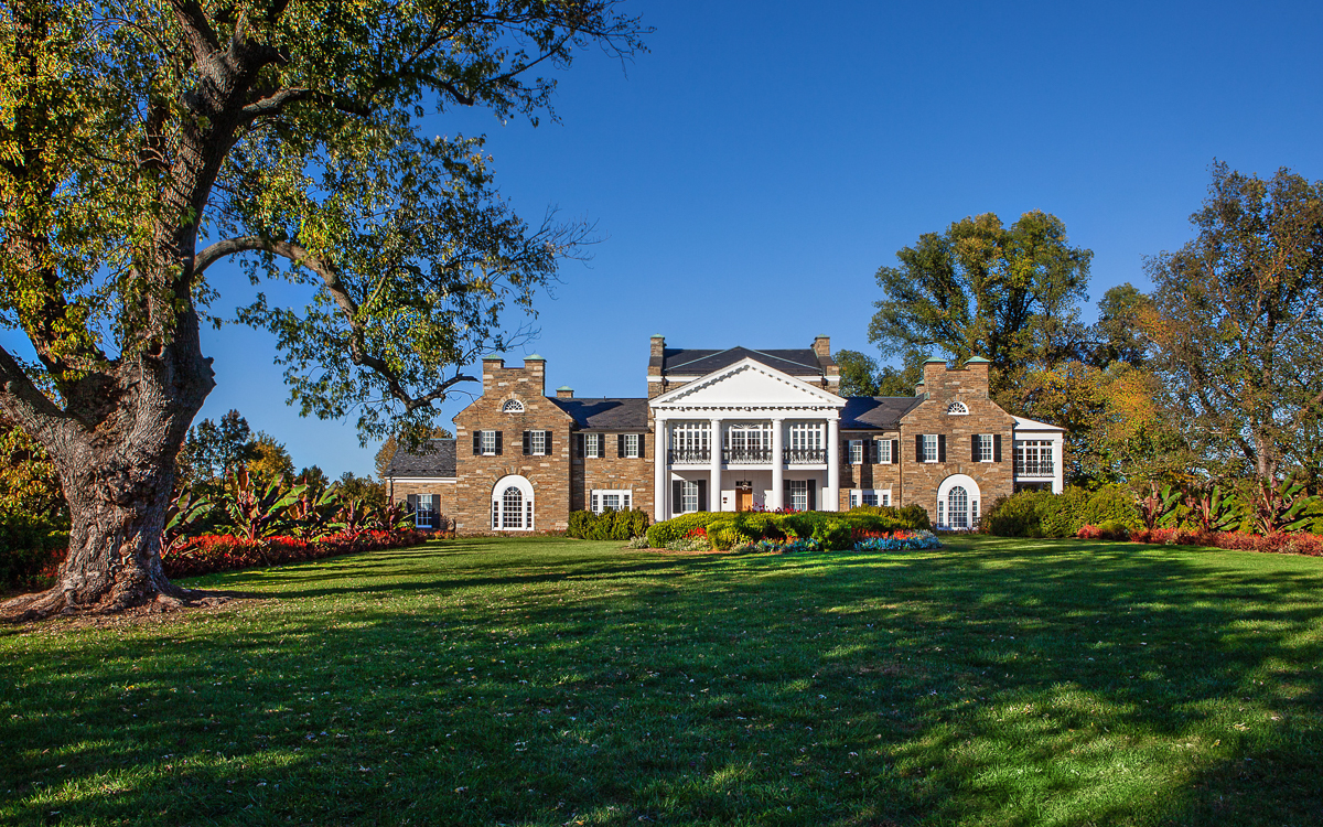 Large two-story mansion with brick exterior, white columns, and multiple chimneys, set on a landscaped lawn with trees and colorful flowerbeds under a clear blue sky.
