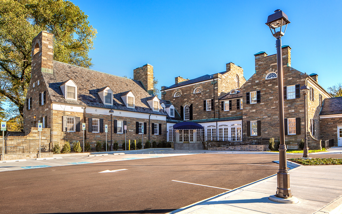 Stone building with multiple chimneys, arched windows, and a slate roof, seen on a clear day with an empty parking lot in the foreground.
