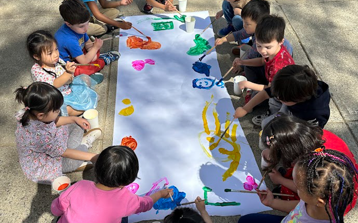 A group of young children sit in a circle on the ground, collaboratively painting colorful designs on a large sheet of white paper outdoors.