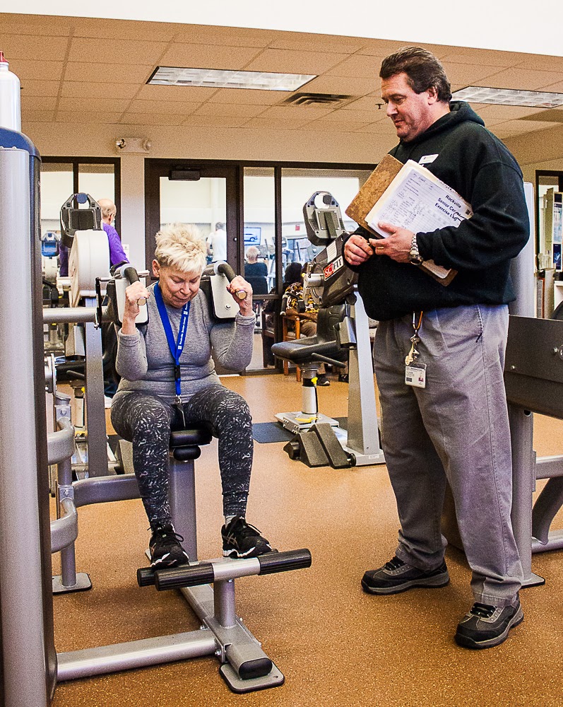An older woman uses a weight machine at a gym while a male trainer stands beside her holding a clipboard and observing.