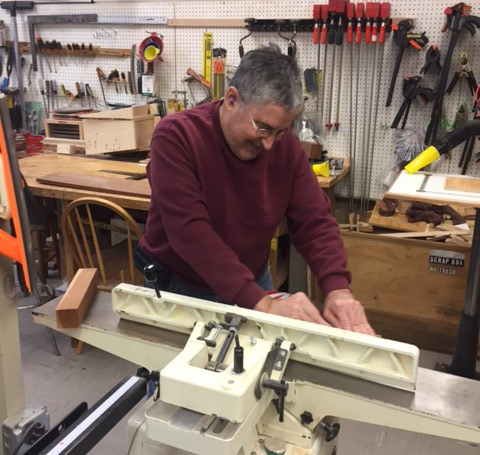 A man wearing a maroon sweater operates a jointer in a woodworking shop filled with tools and equipment.