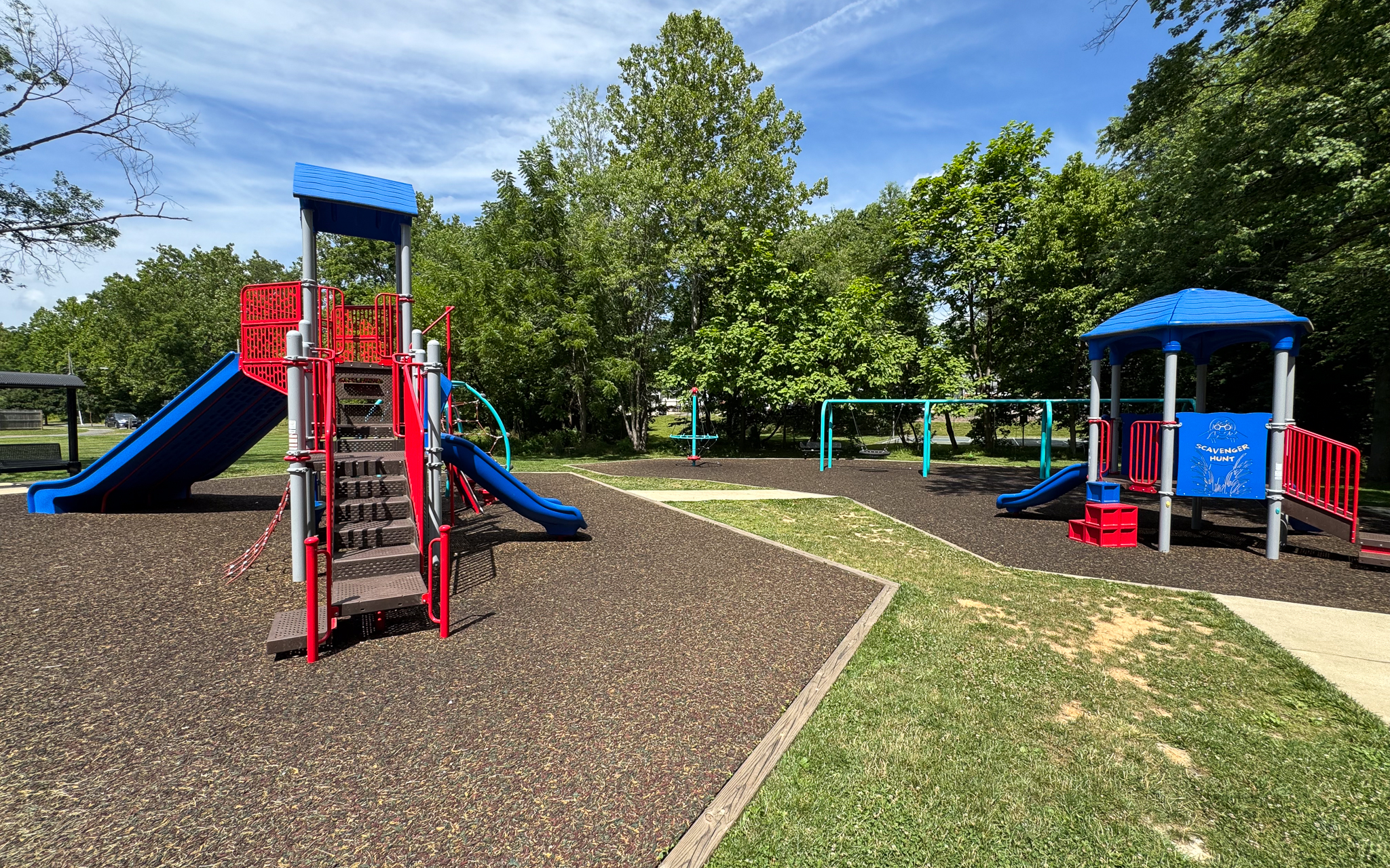 A playground with red and blue climbing structures, slides, swings, and surrounding trees under a sunny sky.