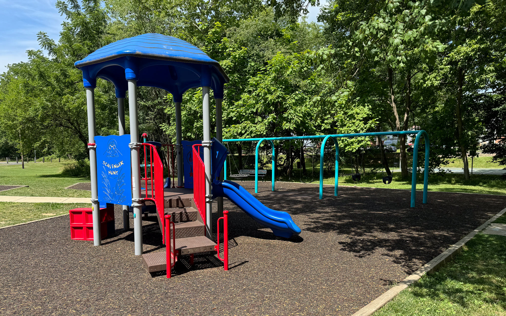 A playground with a blue and red play structure, slide, and swings is surrounded by trees and green grass on a sunny day.