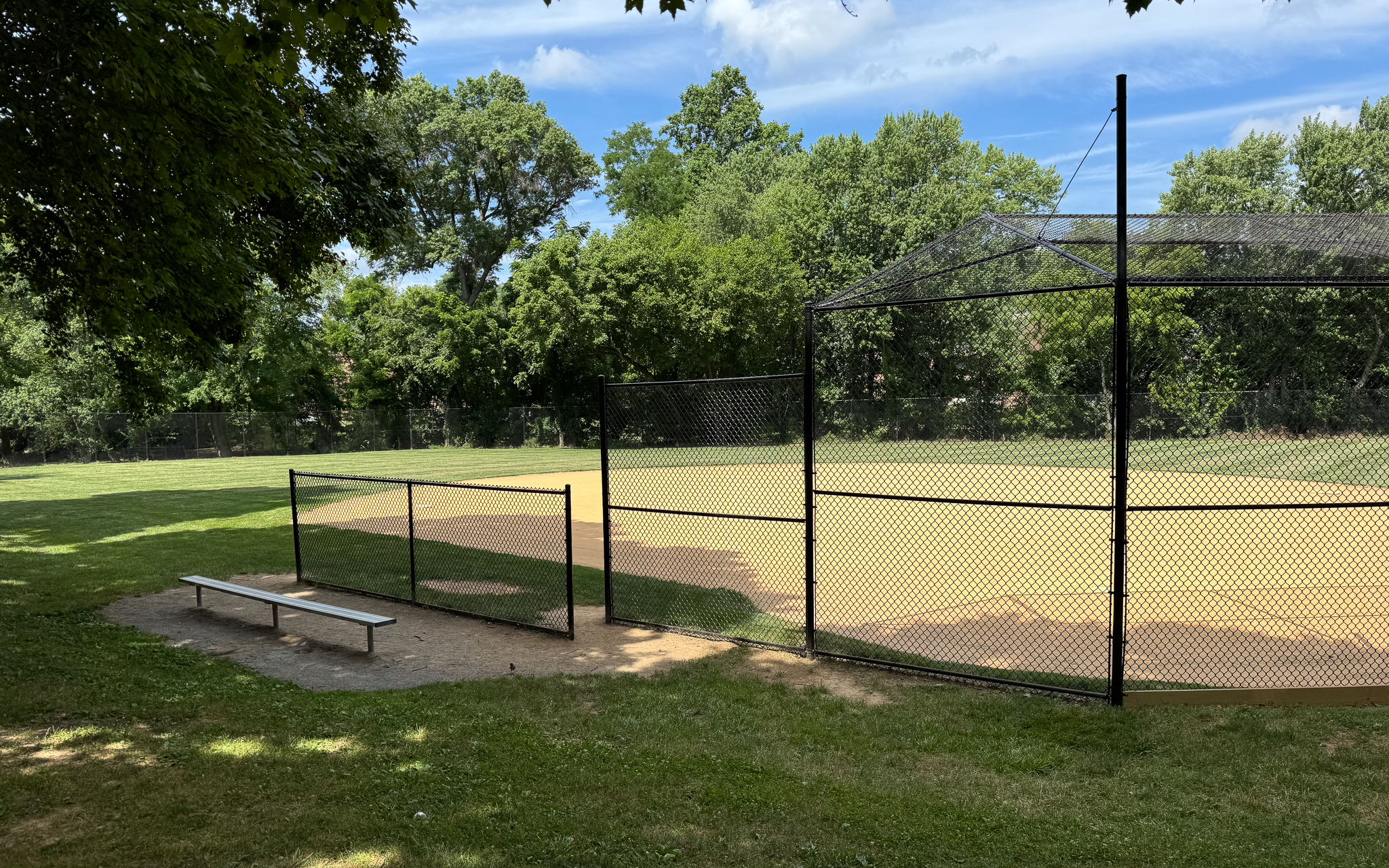 A baseball field with a dirt infield, chain-link backstop, metal bench, and green trees in the background on a sunny day.