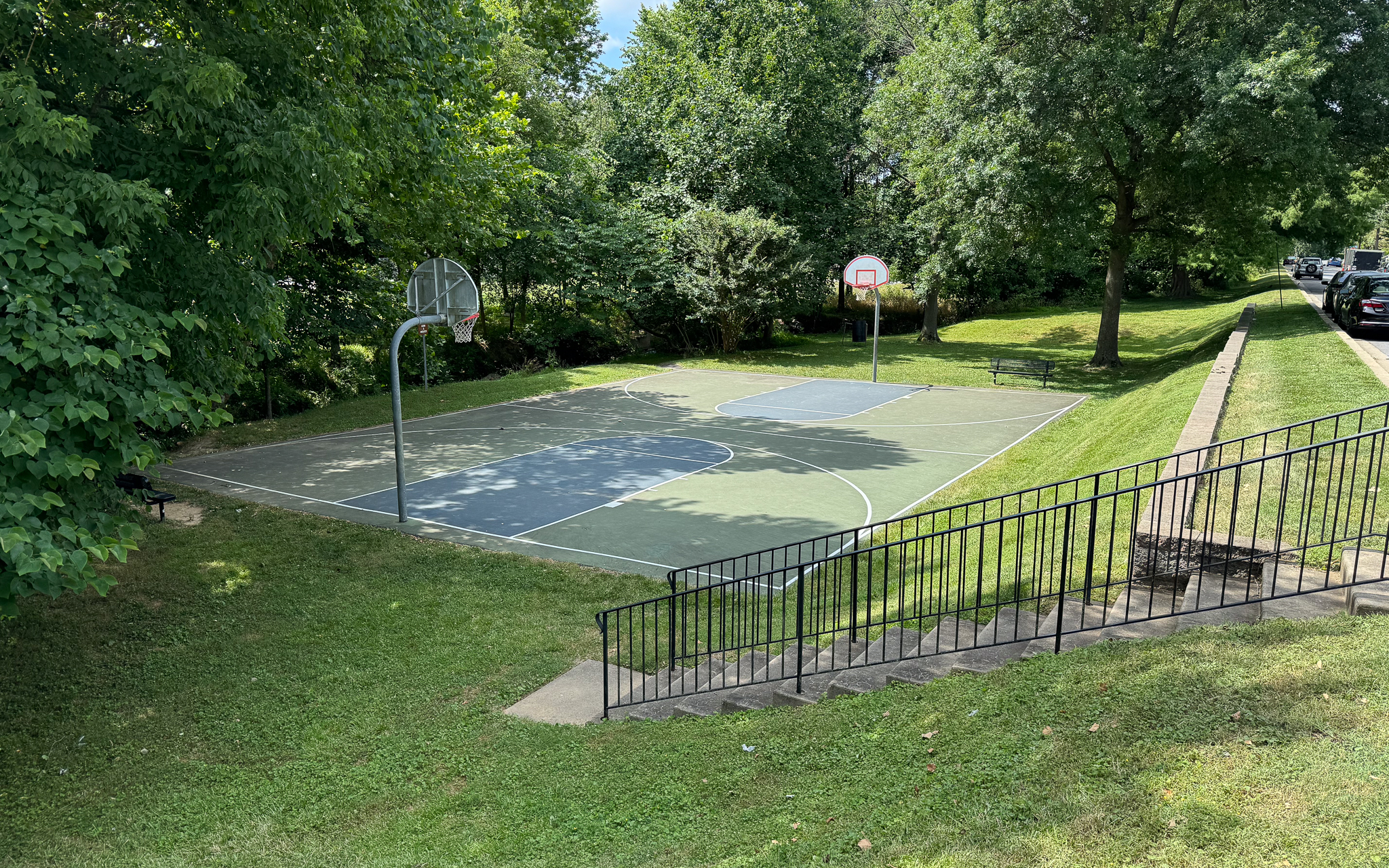 An outdoor basketball court with two hoops, surrounded by grass, trees, and a metal railing along a nearby sidewalk.