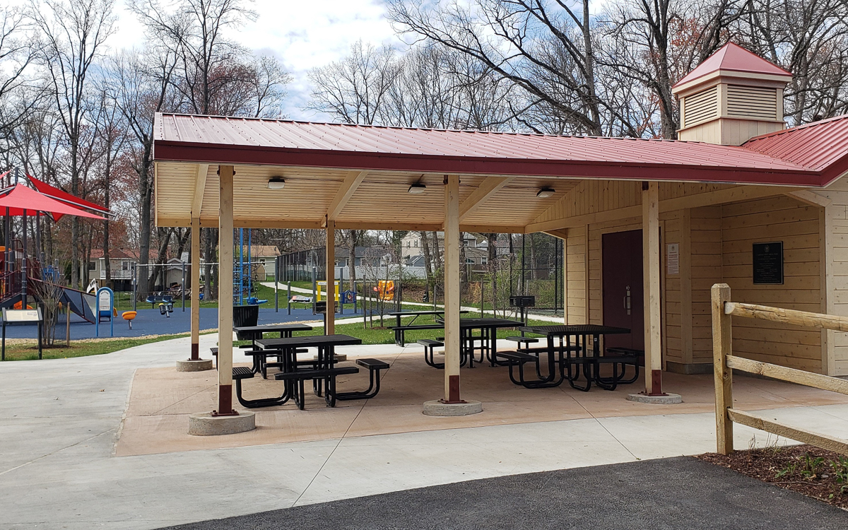 A covered picnic area with black tables next to a playground and sports court in a park, surrounded by trees and houses in the background.
