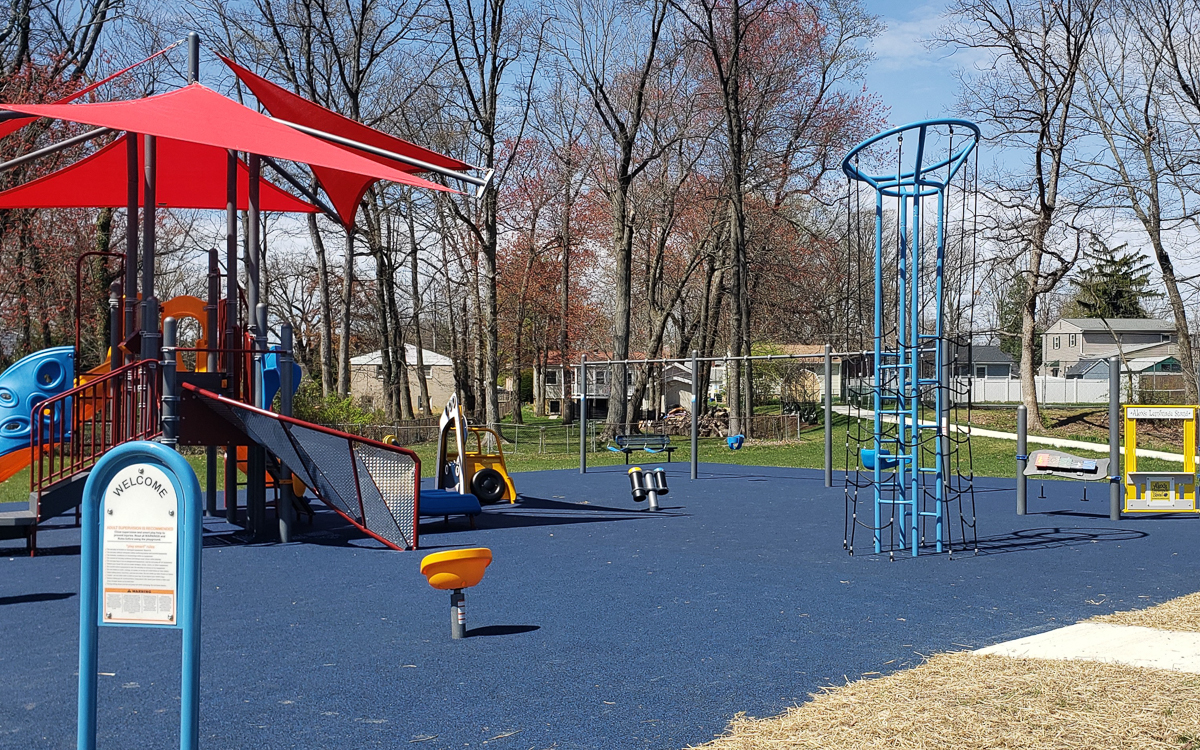 A playground with a blue rubber surface, red canopy, slides, climbing structures, and swings, surrounded by trees and houses in the background.