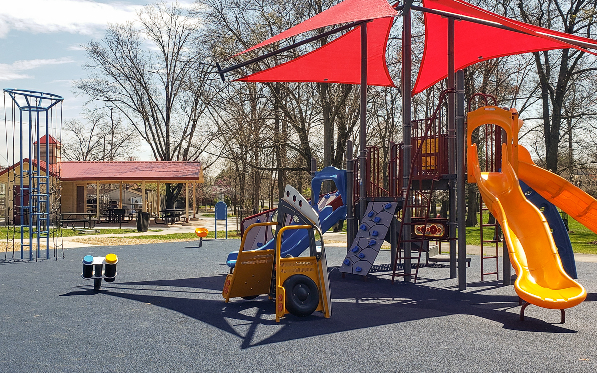 A playground with slides, climbing structures, a play vehicle, and a shaded canopy; trees and a picnic shelter are visible in the background.