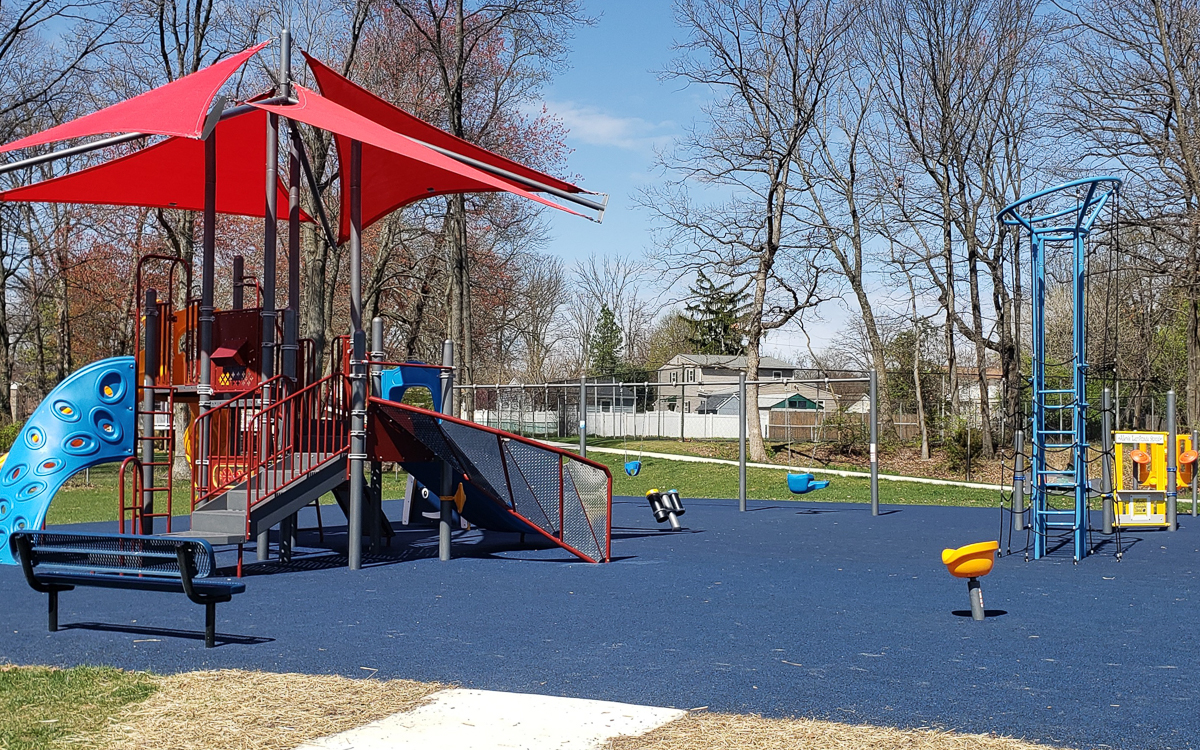 Playground with red canopy, slides, climbing structures, swings, and benches on blue rubber flooring, surrounded by trees and a fence on a sunny day.