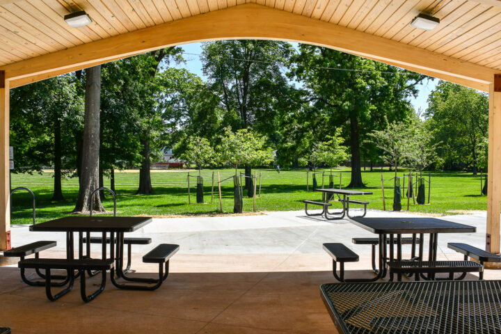 Covered picnic area with metal tables and benches overlooking a green park with trees and a sidewalk on a sunny day.
