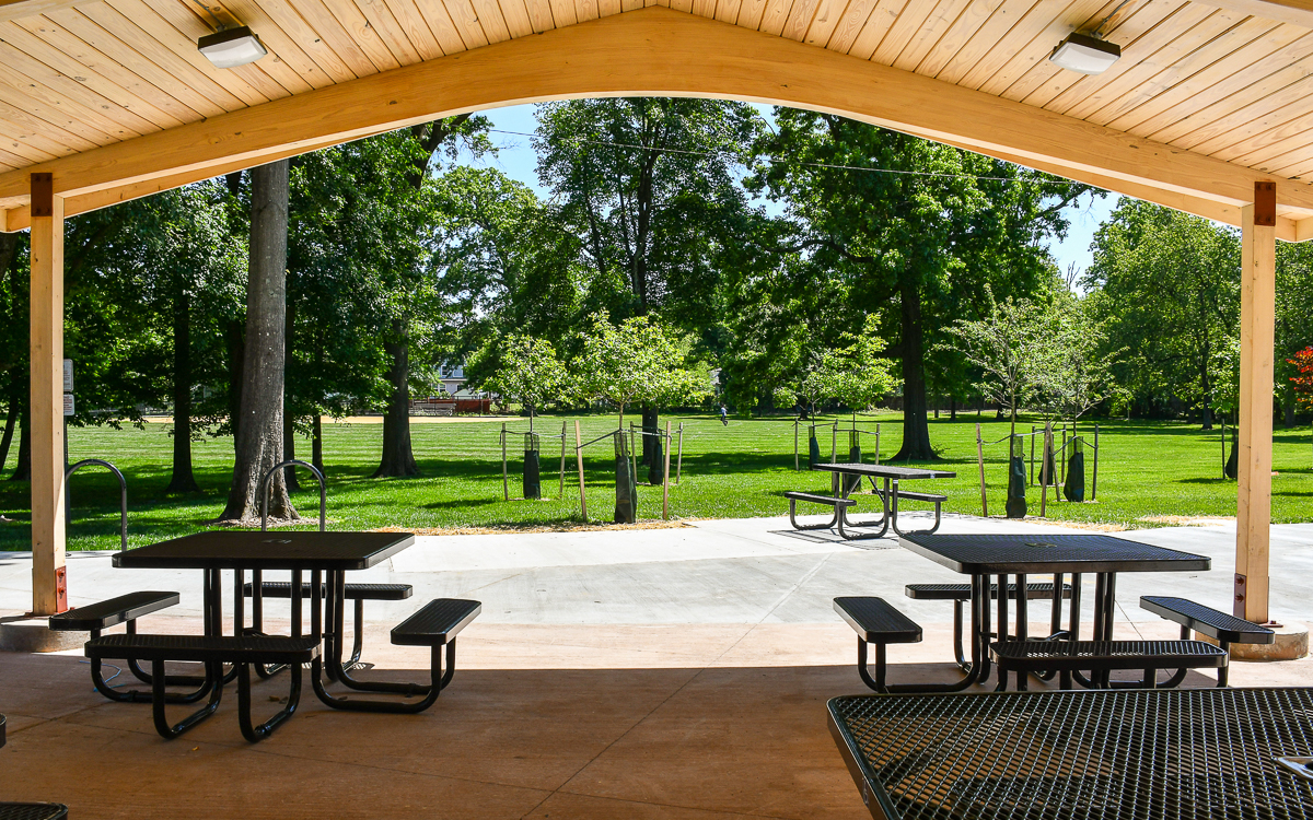 Covered picnic area with metal tables and benches overlooking a green park with trees and a sidewalk on a sunny day.