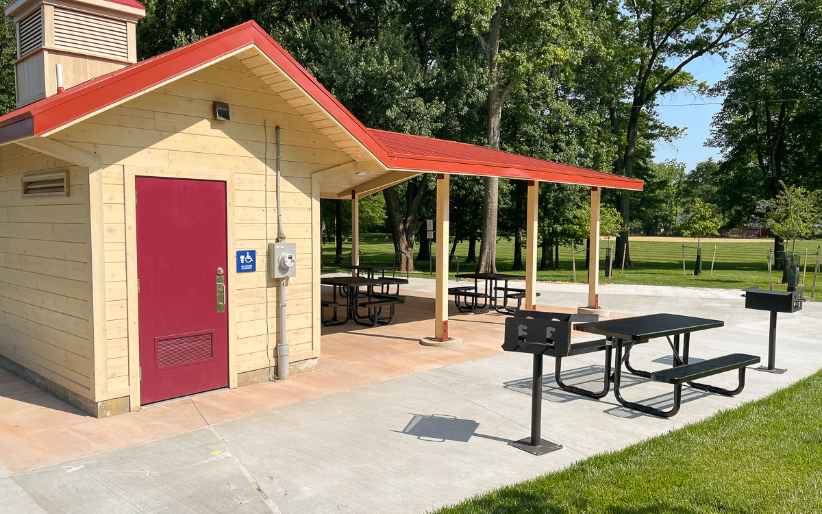 A park shelter with a red roof, picnic tables, grills, and a restroom building, surrounded by grass and trees on a sunny day.
