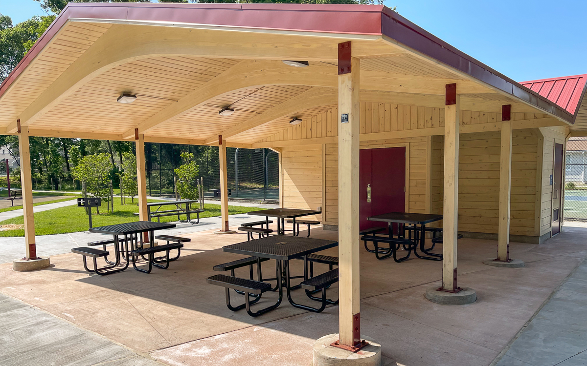 A covered wooden pavilion with picnic tables on a concrete pad in a park, with a red door, nearby grill, and fencing in the background.