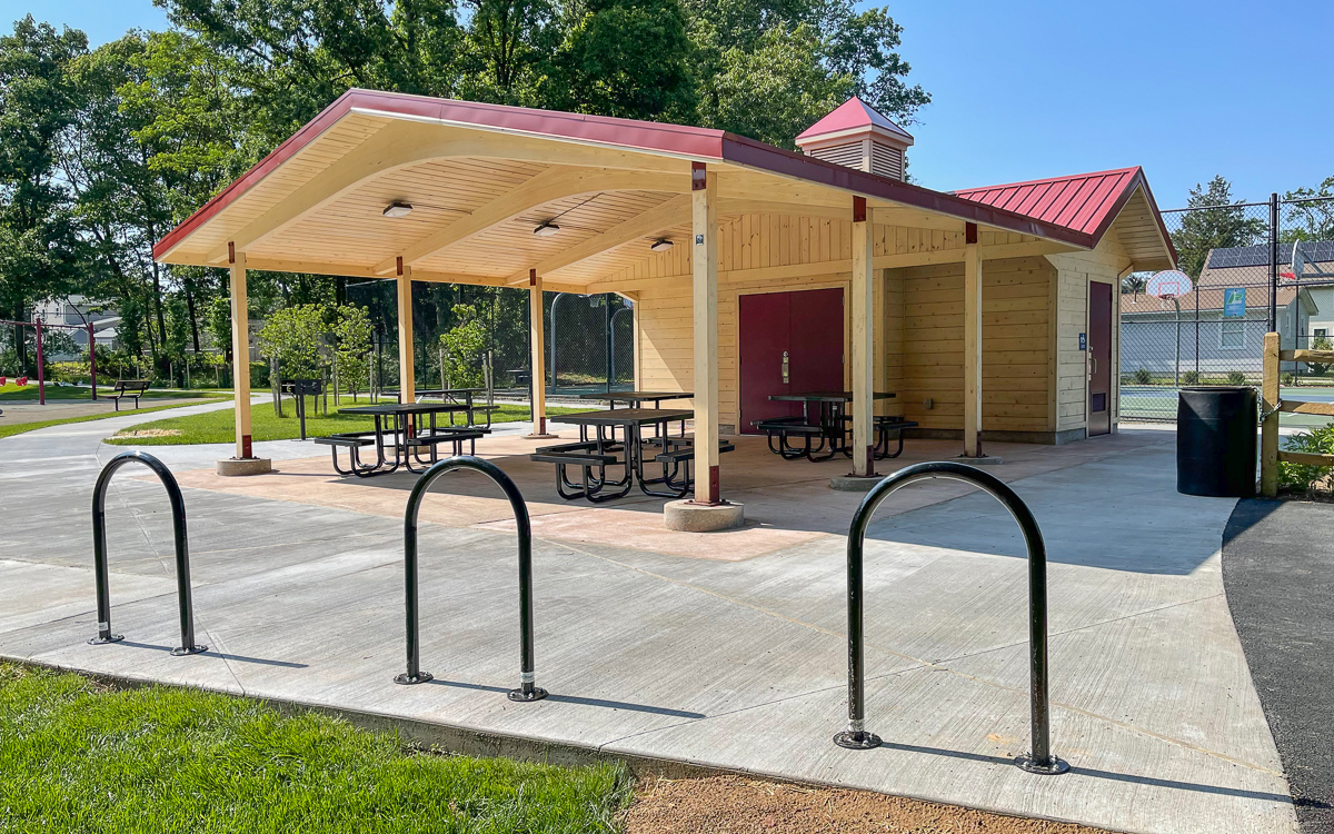 A wooden picnic pavilion with tables and benches sits on a concrete pad beside bike racks, near a tennis court and surrounded by trees on a sunny day.