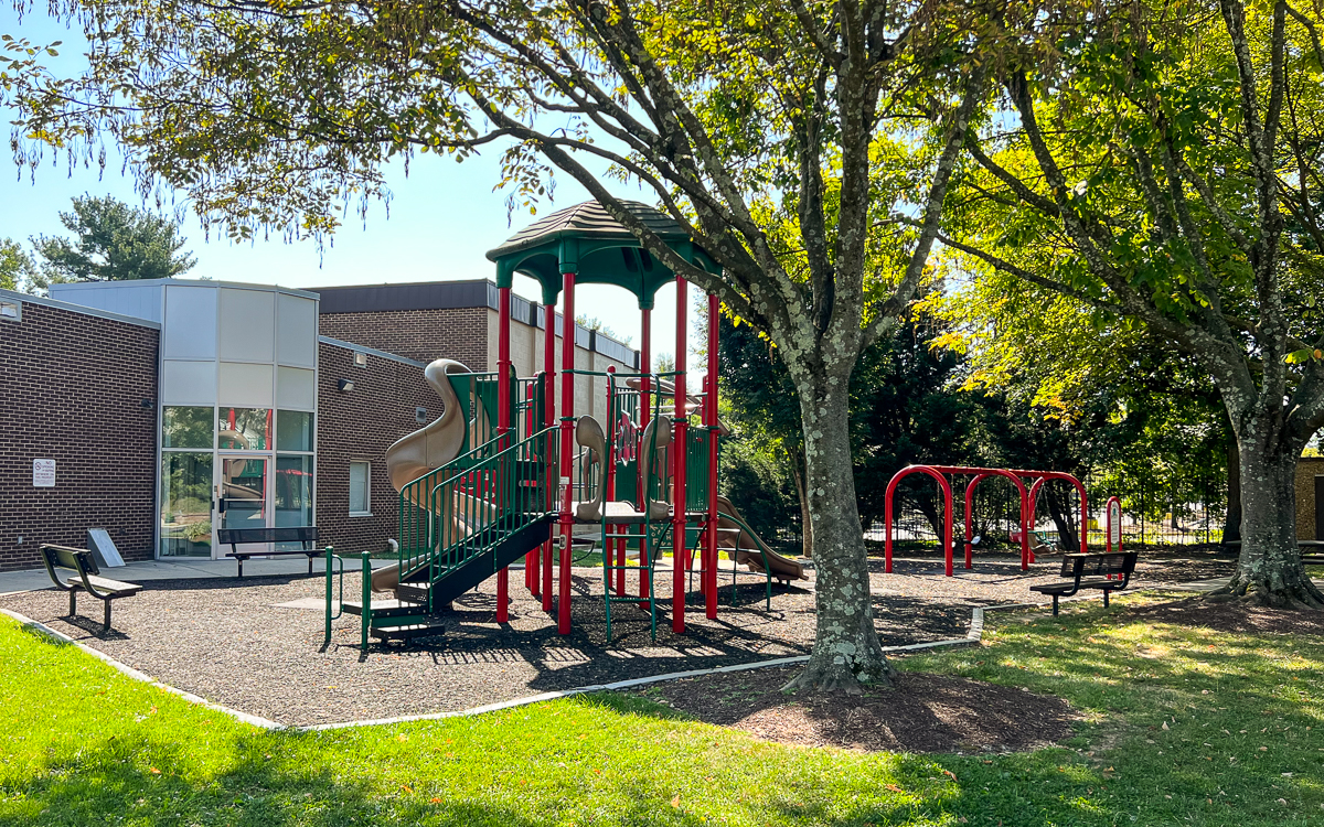 A playground with a slide, climbing structure, and swings is next to a brick building, with benches and trees providing shade on a sunny day.