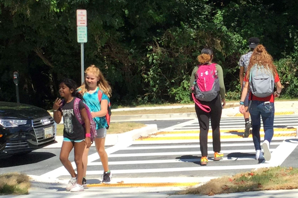 Four children with backpacks walk across a crosswalk in daylight, two heading toward the camera and two walking away, with trees and parked cars in the background.