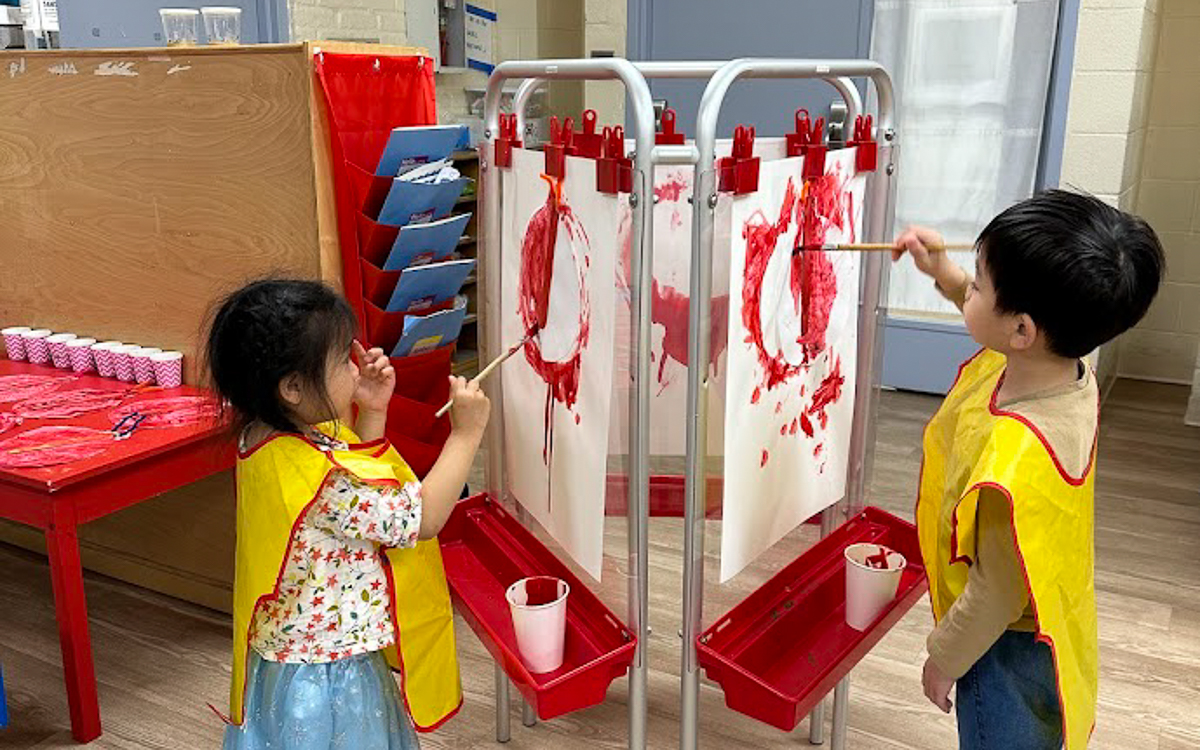 Two young children wearing yellow aprons use paintbrushes to paint on paper mounted on easels. Red paint cups and finished paintings are visible nearby.