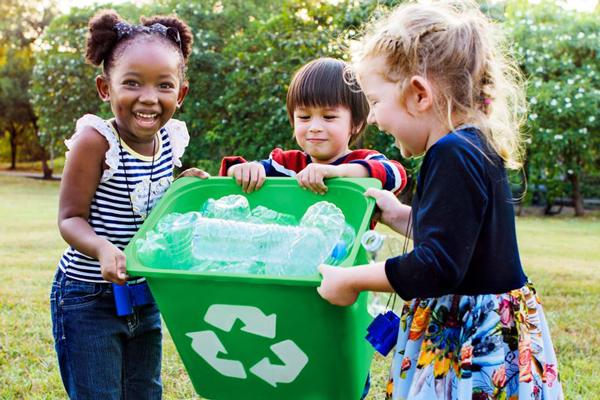 Three children hold a green recycling bin filled with plastic bottles outdoors in a grassy area.