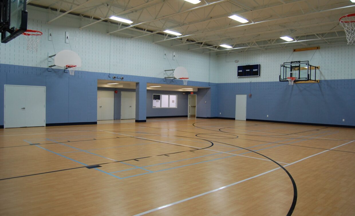 Indoor gymnasium with a light wood floor, several basketball hoops, blue and white walls, and overhead fluorescent lighting. The gym is empty.