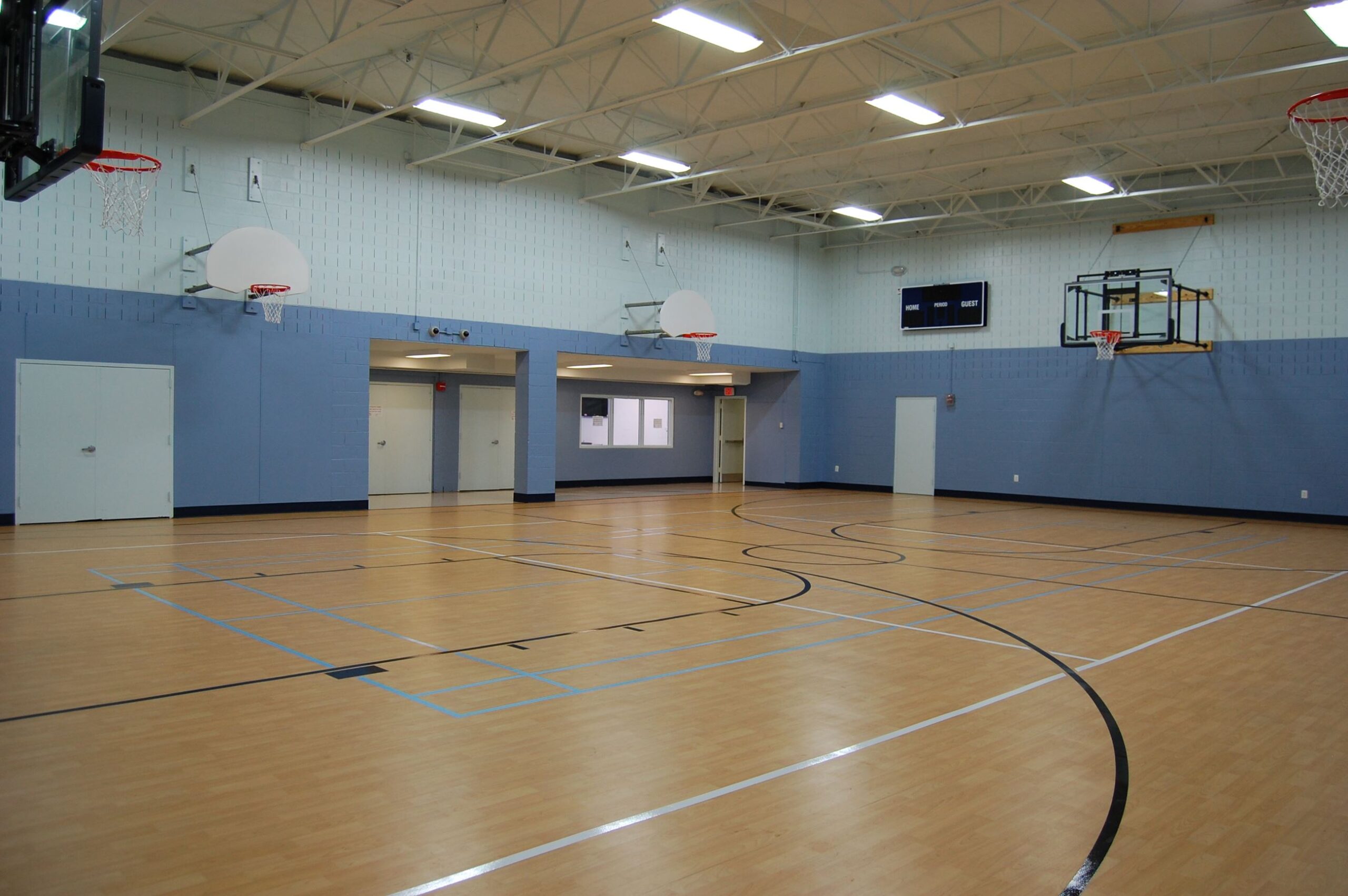 Indoor gymnasium with a light wood floor, several basketball hoops, blue and white walls, and overhead fluorescent lighting. The gym is empty.