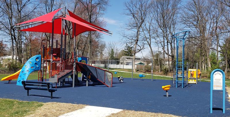 Playground with red-roofed climbing structure, slides, benches, and various play equipment on a blue rubber surface, surrounded by bare trees under a clear sky.